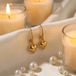 Gold puffed Heart charm earrings on a lever back base, displayed hooked on silk material with pearls and candles in the background